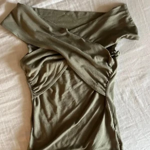 topp - Snygg olivgrön topp med omlott-detalj och offshoulder-design. Toppen är tight och har en croppad passform som framhäver midjan. Tillverkad i mjukt stretchigt material, perfekt för dig som gillar trendiga och unika plagg.