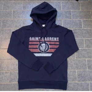 Mörkblå Saint Laurent hoodie med tryck - Snygg mörkblå hoodie från Saint Laurent med huva och snörning. Framsidan har ett coolt tryck med röda och vita ränder samt logga i mitten. Tröjan har långa ärmar och ribbade muddar. Perfekt för dig som gillar streetwear och vill sticka ut. Helt ny, säljer du den är för liten