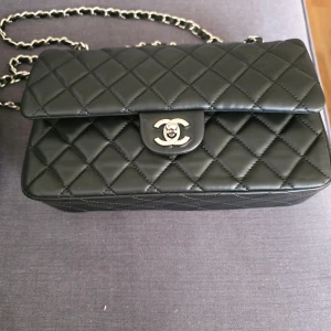 Svart quiltad axelväska i skinn - Svart axelväska från quiltat skinn med silverfärgad kedja och klassiskt CC-spänne. Insidan är vinröd med präglad logga. Perfekt storlek för det viktigaste och en riktig statement piece för din outfit.