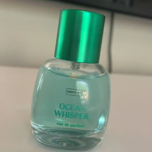 Ocean Whisper Eau de Parfum Sence - Fräsch parfym med havsinspirerad känsla från Sence. Flaskan är rundad och genomskinlig med ljusblå vätska och har ett metallicgrönt lock och spraymunstycke. Perfekt för dig som gillar fräscha och lätta dofter. 