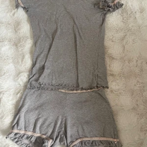 Grå volangset från Intimissimi - Mjuka grått set från Intimissimi med volangkant och ljusrosa spetsdetalj längs bensluten. Shortsen har en relaxed fit och är perfekta för chill dagar hemma. Tillverkade i ett stretchigt bomullsmaterial som känns skönt mot huden.