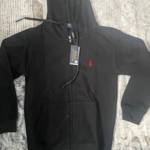 Svart hoodie från Polo Ralph Lauren - Svart hoodie från Polo Ralph Lauren med dragkedja och huva. Klassisk röd broderad logga på bröstet och känguruficka framtill. Tillverkad i mjuk bomull, perfekt för chill dagar. Snygg och enkel att matcha med allt.