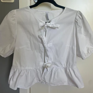 Vit blus med knytning från Stradivarius - Supersöt vit blus från Stradivarius med tre knytrosetter framtill, puffiga korta ärmar och volang nertill. Står att det är storlek M, men skulle säga att den passar nån som i vanliga fall bär XS.