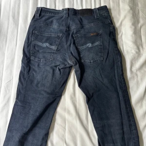 Nudie Jeans Thin Finn blå jeans 32/32 - Snygga blå jeans från Nudie Jeans, modell Thin Finn. Jeansen har klassisk femficksdesign, broderade bakfickor och är tillverkade i 98% ekologisk bomull och 2% elastan för en bekväm passform. Smalare ben och normal midja. Lagad i skrevet men är ingen stor skada och lagningen var förebyggande för hål.