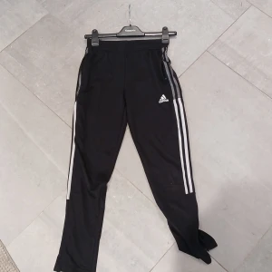 Svarta Adidas träningsbyxor storlek 152 - Svarta träningsbyxor från Adidas med klassiska vita ränder längs sidorna och logga på låret. Byxorna har resår i midjan och dragkedjor vid fickorna. Perfekta för sport och aktivitet. Skriv vid mista funderingar, priset är diskuterbart.