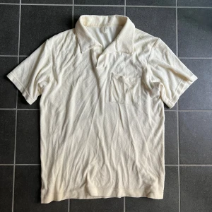 Krämfärgad frotté pike Arket - Krämfärgad piké i frotté från Arket med klassisk krage och bröstficka. Modellen är kortärmad och har en relaxed vibe, perfekt för chill dagar. Materialet är mjukt och ger en lite retro känsla. Enkel och stilren design utan tryck eller mönster. Lite missfärgad i nacken men går säkert bort efter en tvätt, samt inget som syns vid användning!