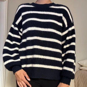 Randig stickad tröja i marinblått och vitt - Mysig stickad tröja med breda marinblå och vita ränder. Tröjan har en avslappnad passform, rund halsringning och långa ärmar med ribbade muddar. 