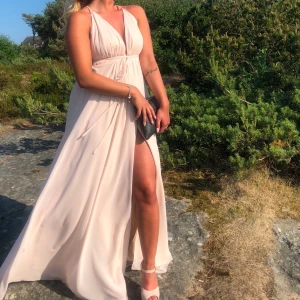 Ljusbeige långklänning med slits - Elegant ljusbeige långklänning i flowigt material med djup v-ringning, smala axelband och hög slits framtill. Klänningen har omlottkänsla och knyt i midjan för snygg passform. Perfekt för sommarens speciella tillfällen. Fläckar finns se bilder, köpt för 2000kr envänd på min bal.