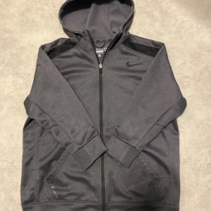 Nike grå hoodie Therma-Fit M - Mörkgrå hoodie från Nike med Therma-Fit-teknologi. Tröjan har huva, hel dragkedja och två fickor framtill. Perfekt för kyliga dagar och träning. Klassisk Nike-logga på bröstet och mjukt fleecefoder på insidan. Har några slitningar men inget som direkt syns vid användning. Längd längd 67cm, bredd 58cm, armlängd 40cm