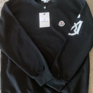 Svart Moncler sweatshirt med logga - Aldrig använd. Svart sweatshirt från Moncler med rund hals, ribbade muddar och stor vit Moncler-logga på ena ärmen. Klassisk broderad Moncler-patch på bröstet. Mjuk och skön bomullskvalitet, perfekt för en clean och stilren look.