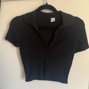 Svart ribbad kortärmad blus med krage - Säljer en svart ribbad blus med korta ärmar och klassisk krage. Toppen har knappar framtill och är croppad i modellen, vilket ger en trendig look. Perfekt att styla med jeans eller kjol för en snygg outfit.