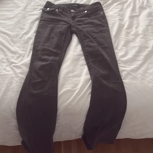 True Religion Joey Low Rise Flare Jeans - Säljer ett par svarta True Religion Joey jeans med låg midja och flare-ben. Klassiska silverfärgade knappar och detaljerade bakfickor med lock och broderi. Jeansen är i stretchig denim och har en snygg passform som framhäver siluetten.