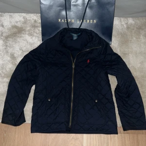 Mörkblå quiltad jacka från Polo Ralph Lauren - Quiltad mörkblå jacka från Polo Ralph Lauren med guldfärgad dragkedja och två fickor med knappstängning. Perfekt höstjacka för dig som clean stil. Priset går att diskutera!