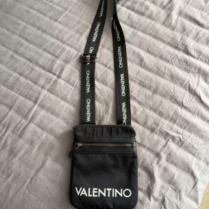 Svart axelväska från Valentino - Snygg svart axelväska från Valentino med justerbart band där loggan står tryckt i vitt. Väskan har en dragkedja framtill och är tillverkad i ett slätt syntetmaterial. Perfekt storlek för mobil och småprylar, enkel att bära crossbody.