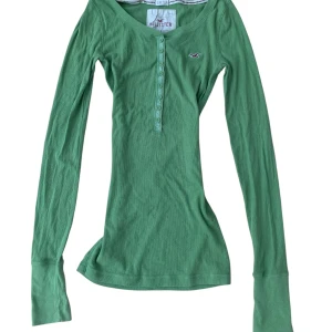 Grön långärmad Henley tröja från Hollister - Stretchig grön långärmad tröja från Hollister med rund halsringning och knappslå framtill. Tröjan har en liten broderad logga på bröstet och är tillverkad i en mjuk bomulls- och polyestermix. Perfekt för lager-på-lager och chill dagar.