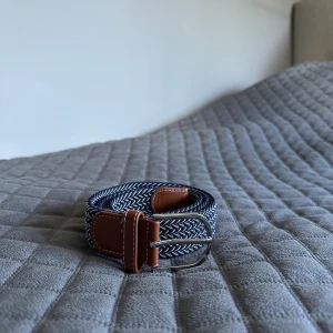 Blåttvitt flätat bälte med skinn-detaljer - Snyggt flätat bälte i marinblått och vitt med bruna skinn-detaljer och silverfärgat spänne. Bältet har ett coolt mönster och passar perfekt till jeans eller chinos. Stilren design som lyfter din outfit.