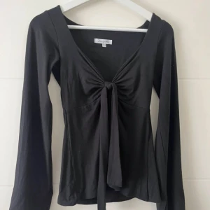 Svart långärmad topp med knyt från DesignSi - Svart långärmad topp från DesignSi med djup v-ringning och snygg knytning framtill. Toppen har en figurnära passform och är tillverkad i mjuk bomull, perfekt att styla med jeans eller kjol för en trendig look.