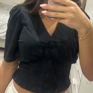Svart blus med stora knappar - Säljer en svart blus med v-ringning och stora klädda knappar framtill. Toppen har korta puffärmar och smockad midja som ger en snygg siluett. Materialet känns mjukt och lätt, perfekt för att lyfta din outfit.