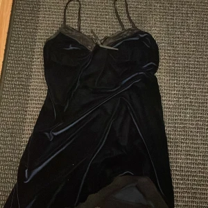 Svart sammetsklänning med spetsdetaljer - Supersnygg svart kortklänning i sammetsliknande polyester med tunna axelband och spetsdetaljer upptill samt liten rosett framtill. Klänningen har en figurnära passform och är perfekt för dig som gillar en elegant och stilren look.