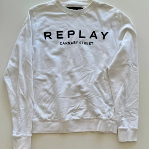 Vit sweatshirt från Replay - Vit sweatshirt från Replay med svart tryck 'REPLAY CARNABY STREET' på bröstet. Klassisk rund halsringning och långa ärmar. Mjuk bomullskvalitet och ribbade muddar vid ärmslut och nederkant. Perfekt för en clean och stilren look.