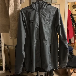 Grå Liverpool FC vindjacka med huva - Säljer en grå vindjacka från Liverpool FC med broderad logga på bröstet. Jackan har huva, hel dragkedja framtill och två sidofickor. Perfekt för dig som vill visa ditt stöd för laget även när det blåser ute. Funkar även för regn står att de är storlek S men är mer som en M/L. Dessutom aldrig använd då den inte passar mig 