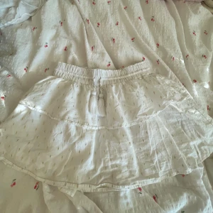 Vit kortkjol med guldtrådar från Gina Tricot - Supersöt vit kortkjol från Gina Tricot med glittriga guldtrådar, volang och elastisk midja med dekorativa tofsar och pärldetaljer. Perfekt för dig som vill ha en lätt och luftig kjol med extra bling.