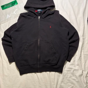 Svart hoodie från Polo Ralph Lauren - Svart hoodie från Polo Ralph Lauren med dragkedja och huva. Klassisk röd broderad logga på bröstet och stora fickor framtill. Tillverkad i mjuk bomull, perfekt för chill dagar. Snygg och enkel att matcha med allt.