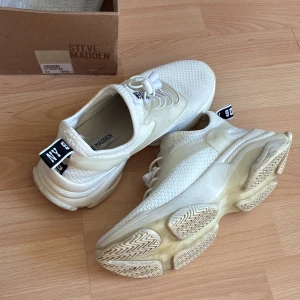 Vita sneakers från Steve Madden - Säljer ett par fräscha vita sneakers från Steve Madden i storlek 37. Skorna har stickad ovandel i syntetmaterial, coola detaljer med transparenta partier och snörning framtill. Tjock sula och svartvit logga på hälen ger en sportig vibe.