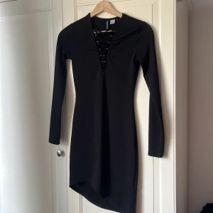 Svart snörad klänning H&M Divided - Svart kortklänning från H&M Divided med lång ärm och snygg snörning framtill i urringningen. Klänningen har en tight passform och är tillverkad i ett stretchigt material som sitter skönt på kroppen. Perfekt för dig som gillar en stilren look med en twist.
