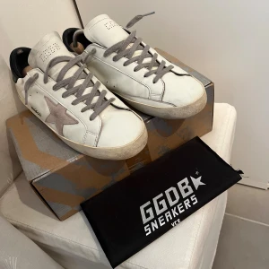 Golden Goose vita sneakers med stjärna - Golden Goose sneakers i vitt skinn med grå mockadetaljer och ikonisk stjärna på sidan. Grå snörning, svart häl och platt sula. Snyggt slitna för en cool streetstyle-look. Kommer med originalkartong och dustbag.