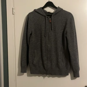 Grå kashmir hoodie från Sayless - Mysig grå hoodie från Sayless i lyxig kashmir. Tröjan har dragkedja framtill, huva med snörning och två fickor. Perfekt för kalla dagar och chill vibes. Ribbade muddar och snygg detalj i läder på dragkedjan. Tröjan är sprillans ny. Köptes i vintras men har inte använt den då den har varit lite för stor och inte min typ.