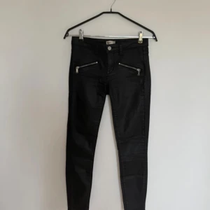 Svarta skinny jeans med dragkedjor - Säljer ett par svarta skinny jeans med coola silvriga dragkedjor framtill. Byxorna har en tight passform och är tillverkade i ett glansigt material som ger en snygg look. Perfekta för dig som gillar en edgy stil och vill sticka ut lite extra.