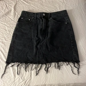 Svart jeanskjol med fransad kant - Cool svart jeanskjol i modell med fem fickor och fransad nederkant. Kjolen är kort och har en klassisk knapp- och dragkedjestängning framtill.🖤