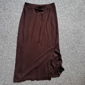 Brun långkjol med slits - Mörkbrun långkjol i mjukt, stretchigt material. Kjolen har en enkel och stilren design med en diskret slits på sidan som ger en snygg detalj. Perfekt att styla med sneakers eller boots för en chill look.