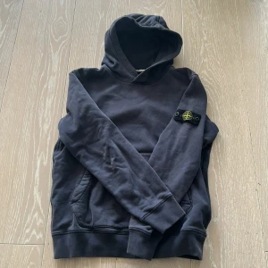 Blå hoodie från Stone Island Junior - Snygg svart hoodie från Stone Island Junior med klassisk logga på ärmen. Tröjan har huva, känguruficka och långa ärmar. Tillverkad i mjuk bomull, perfekt för en chill och stilren look. Tröjan är lite urtvättad, men inte så mycket man tänker på. Jag är 160 cm lång och den sitter bra. 