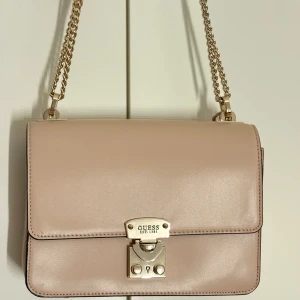 Ljusrosa axelväska från Guess med kedja - Snygg ljusrosa axelväska från Guess med guldfärgad kedjerem och låsdetalj framtill. Väskan har stilren, boxig form och är tillverkad i slätt syntetmaterial. Baksidan har en praktisk dragkedjeficka och ett dekorativt G-hänge i metall. Använd en gång & är i väldigt fint skick. Mycket förvaring & väldigt fint. 