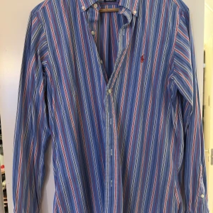 Randig skjorta från Ralph Lauren - Säljer en klassisk blå skjorta från Ralph Lauren med röda och vita ränder. Skjortan har lång ärm, knappar framtill och den ikoniska broderade loggan på bröstet. Modellen är Custom Fit och tillverkad i 100% bomull. Perfekt för dig som gillar preppy stil.