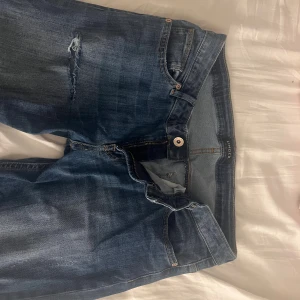 Blå bootcut jeans med slitningar från Lindex - Säljer ett par blå bootcut jeans från Lindex med snygga slitningar och hål vid knäna. Jeansen har klassisk femficksdesign, dragkedja och knapp framtill. Materialet är jeans i bomull och passformen är normal med utsvängda ben.