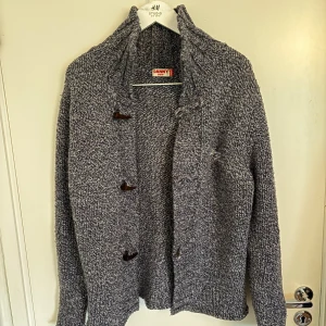 Gråmelerad stickad kofta från Danny - Säljer en gråmelerad stickad kofta från Danny DDW i storlek medium. Koftan har snygga träknappar framtill, hög krage och långärmad design. Perfekt för lager-på-lager och chillare dagar. Grymt mysig och stilren att slänga på sig.