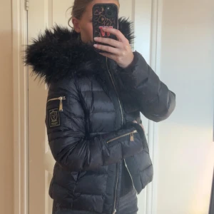 Svart pufferjacka från Chiara Forthi - Säljer en svart pufferjacka från Chiara Forthi med stor fluffig fuskpäls på huvan, guldiga dragkedjor och snygg logga på ärmen. Jackan har quiltad design och är perfekt för kalla dagar. Riktigt stilren och trendig modell.