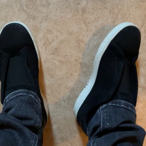 Svarta slip-on sneakers med vit sula - Säljer ett par sköna arigatos skor jag köpt i slutet av sommaren. Har använt dem lite men skulle säga att skicket är på 8,5/10 . Skorna har en minimalistisk design utan snörning och är tillverkade i mocka och syntet. Perfekta för dig som gillar en clean och stilren look. Tveka inte på att höra av er vid pris eller så. Skorna är även i storlek 42 men skulle säga att dem sitter som 43 