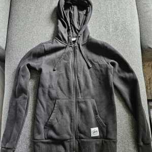 Svart hoodie från Andy by Frank Dandy - Svart hoodie med dragkedja från Andy by Frank Dandy. Tröjan har huva med snörning, två stora fickor framtill och ribbade muddar. Perfekt för kyliga dagar och enkel att matcha med jeans eller joggers.