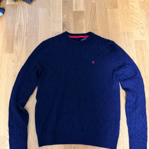 Marinblå stickad tröja från Morris  - Snygg marinblå stickad tröja från morris med klassiskt kabelmönster och rund hals. Tröjan har en liten röd logga broderad på bröstet och ribbade muddar vid ärmslut och nederkant. Perfekt för dig som gillar stilrena och tidlösa plagg.