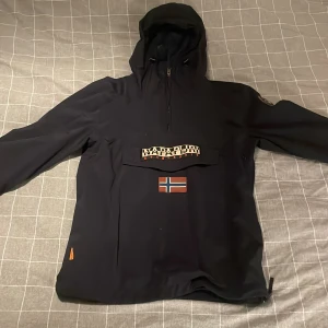 Svart vindjacka från Napapijri - Svart vindjacka från Napapijri med huva, halv dragkedja och stor ficka framtill med logga och norsk flagga. Jackan är tillverkad i slitstark polyester. Perfekt för blåsiga dagar och höst vädret. Den är i storlek L men passar även om du har M. Det är bara att skriva om du har några frågor.