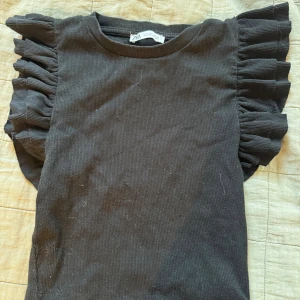 Svart volangärmad topp från Zara - Supersnygg svart topp från Zara med volangdetaljer på axlarna som ger en cool och trendig look. Toppen är ribbad och har en rund halsringning. Perfekt för dig som vill sticka ut lite extra med dina outfits.