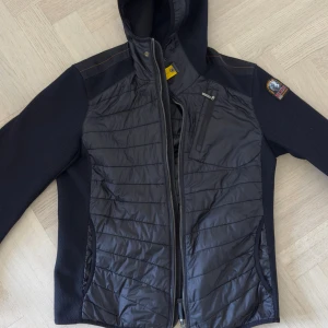 Svart hybridjacka från Parajumpers - Snygg svart jacka från Parajumpers med quiltad front och ärmar i stickad struktur. Jackan har huva, dragkedja och en gul detalj vid halsen. Märkespatch på ena ärmen. Perfekt för kyliga dagar.