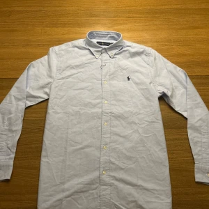 Ljusblå skjorta från Ralph Lauren - Snygg ljusblå skjorta från Ralph Lauren med klassisk button-down krage och broderad logga på bröstet. Skjortan har långa ärmar, vita knappar och är gjord i mjuk bomull. Perfekt för dig som gillar stilrena och tidlösa plagg.