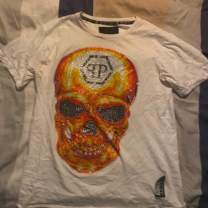 Vit Philipp Plein t-shirt med dödskalle - Snygg vit t-shirt från Philipp Plein med stor dödskalle framtill i orange, gul och svart med glittriga detaljer. Klassisk passform och korta ärmar. Märkeslapp nertill och logga i nacken. Perfekt för dig som gillar statement-plagg.