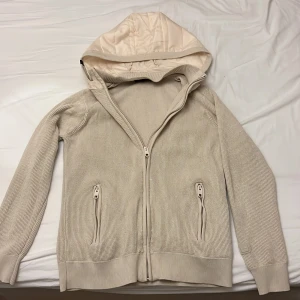 Beige stickad cardigan från Indicode - Säljer en beige stickad cardigan från Indicode i storlek M. Tröjan har dragkedja framtill, två fickor med dragkedja och en fodrad huva. Använd sparsamt, skick 8/10. Passar dig som har mellan s-m!