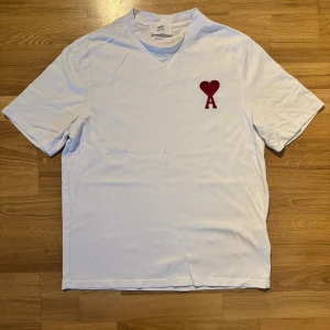 Vit t-shirt från AMI med rött hjärta - Vit t-shirt från AMI med rött hjärta och bokstaven A broderat på bröstet. Klassisk passform, rund hals och korta ärmar. Materialet är mjuk bomull och logotypen AMI är broderad i nacken. Perfekt basic med snygg detalj.